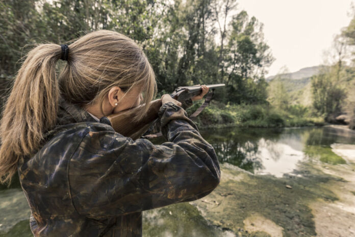 Eine Frau mit einer braun grünen Jacke und einem Zopf ist von hinten zu sehen, während sie mit einem Gewehr im Wald steht. Die Waffe ist auf ein Tier gerichtet, welches sie jagen möchte.