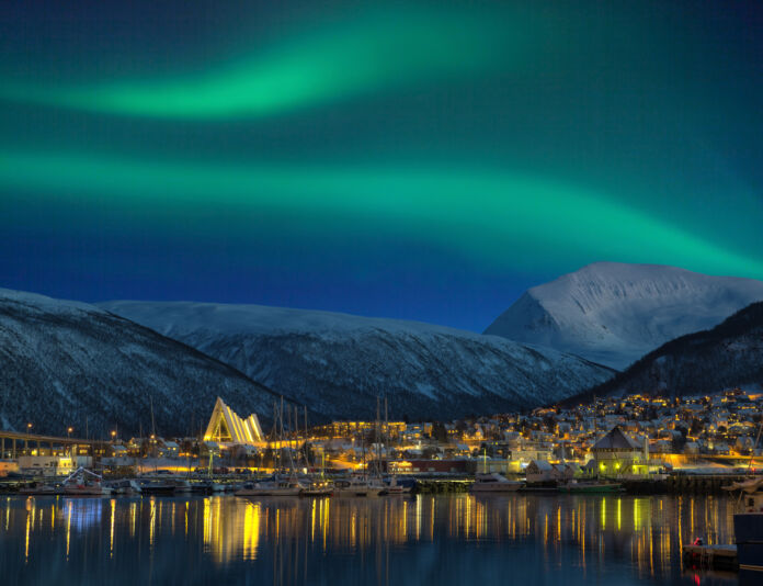 Grüne Polarlichter sind am Nachthimmel über einer kleinen Stadt mit beleuchteten Häusern und einer Kathedrale zu sehen. Die Berglandschaft, das kleine Dorf und das Wasser werden durch die Aurora beleuchtet.