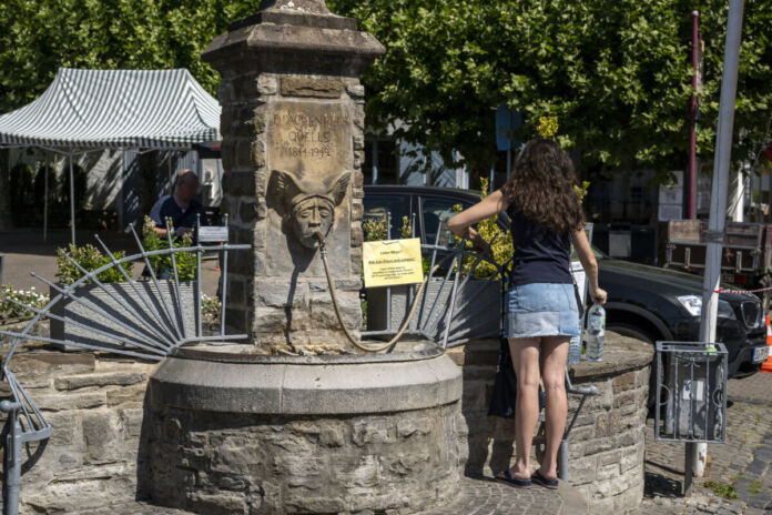 Eine Frau steht vor einem Trinkwasserbrunnen und füllt sich kostenlos ihre Flaschen. Es ist ein heißer, sonniger Tag, das Trinkwasser an diesem Brunnen kostet nichts.