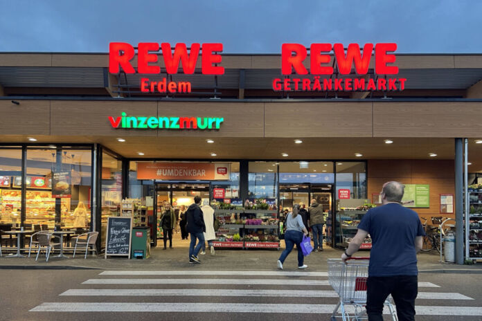 Ein beleuchteter riesiger Eingang eines Rewe Supermarktes mit einem Getränkemarkt von Rewe daneben. Vor der Filiale gehen einige Kunden über den Zebrastreifen und betreten den Supermarkt. Links befindet sich eine Bäckerei.