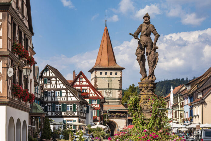 Die steinerne Statue eines Ritters in Rüstung steht in einer charmanten Altstadt vor Fachwerkhäusern und begeisterten Touristen, welche die Gegend erkunden.