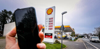 Eine Hand hält im Vordergrund des Bildes ein Handy. Im Hintergrund ist eine Shell-Tankstelle. Autos halten und tanken, während der Fokus auf dem schwarzen Smartphone liegt. Die Handynutzung an Tankstellen ist mit absoluter Vorsicht zu genießen.