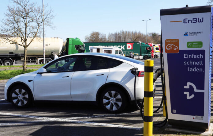 Ein E-Auto wird auf einem Parkplatz an einer E-Ladestation mit einem Kabel aufgeladen. Im Hintergrund befinden sich einige Lkw, die ebenfalls auf dem Parkplatz stehen.