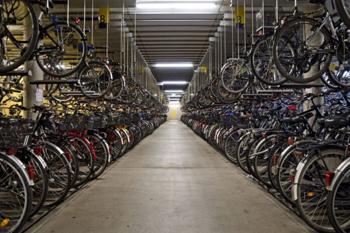 Große beleuchtete Lagerhalle mit mehreren Hundert abgestellten Fahrrädern in einer Reihe links und rechts. Darüber hängen und parken Fahrräder in zweiter Reihe.