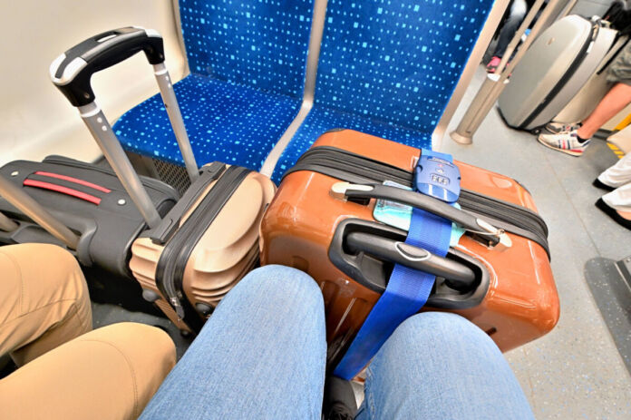Zwei Menschen sitzen in einem Zug auf einem Vierer-Platz und haben vor ihren Knien drei Koffer stehen. An einer Tür wartet eine Schlange von Menschen, bis der Zug hält und sie aussteigen können.