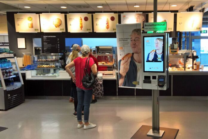 Kunden stehen vor einem Bedienungsterminal an einer IKEA-Theke. In dem schwedischen Möbelhaus werden verschiedene Gerichte wie Hot Dogs und Pommes verkauft.