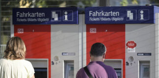 Zwei Menschen stehen an einem Bahnhof nebeneinander an zwei verschiedenen Fahrkarten-Automaten an einem Bahnhof, um ein Ticket für die Fahrt mit der Deutschen Bahn zu lösen.