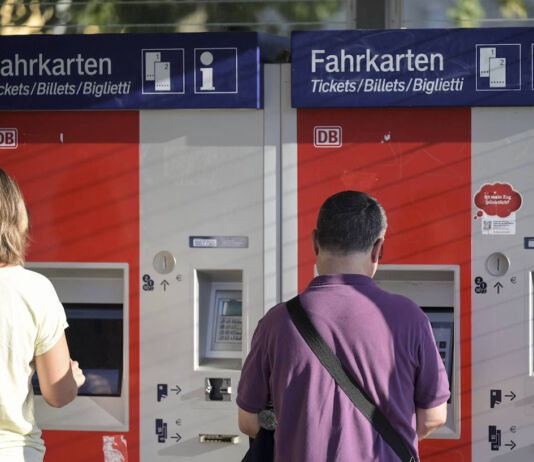 Zwei Menschen stehen an einem Bahnhof nebeneinander an zwei verschiedenen Fahrkarten-Automaten an einem Bahnhof, um ein Ticket für die Fahrt mit der Deutschen Bahn zu lösen.