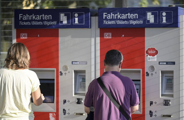 Zwei Menschen stehen an einem Bahnhof nebeneinander an zwei verschiedenen Fahrkarten-Automaten an einem Bahnhof, um ein Ticket für die Fahrt mit der Deutschen Bahn zu lösen.