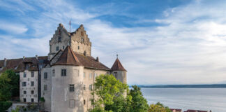 Eine alte, noch bewohnte Burg ragt zwischen Bäumen und Häusern hervor. Im Hintergrund ist ein großer See. Der Horizont ist weit, der Himmel ist blau. Die Festung ist aus Stein, auf dem Dach prangt eine Fahne.