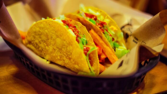 Kross gebackene und lecker gefüllte Tacos stehen auf einem Servierkorb. Unter ihnen liegt eine Serviette. Die Farbtöne sind in gelben Nuancen gehalten.