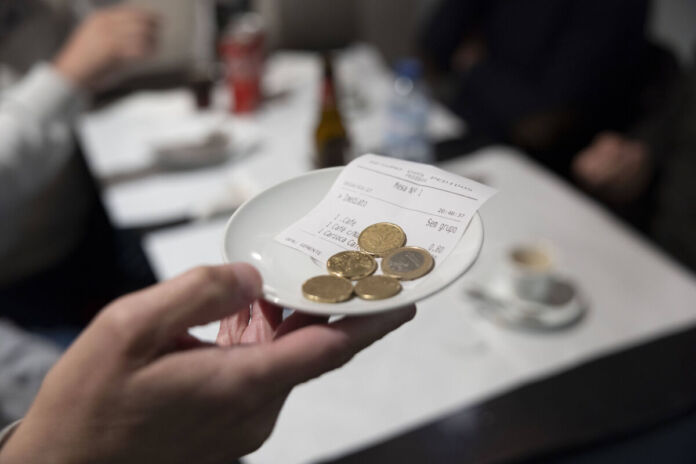 Eine Person hält ein kleines Schälchen in der Hand. In der Schale liegen Euro-Münzen. Im Hintergrund ist ein Tisch in einem Restaurant zu sehen.