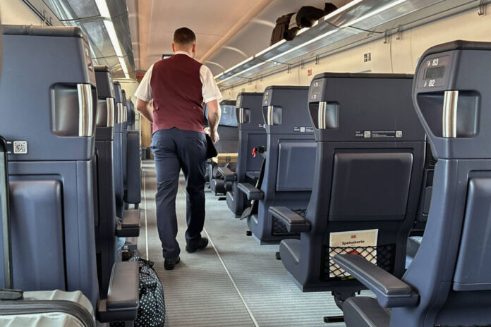 Ein Zugbegleiter der Deutschen Bahn trägt seine Arbeitskleidung und geht durch die Reihen eines Zugabteils. Ein Rucksack befindet sich auf der Ablage über den Sitzen.