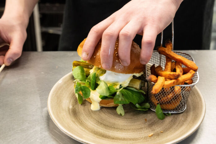 Die Hände eines Kochs sind gerade dabei, einen Burger mit Spargel zuzubereiten. Dieser liegt auf einem Teller. Daneben befindet sich ein Gitterkorb mit Pommes. Die Restaurants eines beliebten Promis sind nun insolvent.