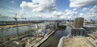 Die Luftaufnahme der eines Hafens bei blauem Himmel. Eine Baustelle am Hafen mit Kränen zwischen hohen Gebäuden, Autos und Schiffen.