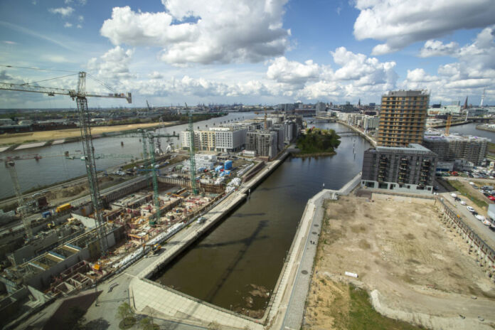 Die Luftaufnahme der eines Hafens bei blauem Himmel. Eine Baustelle am Hafen mit Kränen zwischen hohen Gebäuden, Autos und Schiffen.