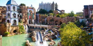 Die Achterbahn Chiapas im Phantasialand fährt durch das Wasser. Im Hintergrund sind weitere Fahrgeschäfte zu sehen. Es scheint Sommer zu sein.