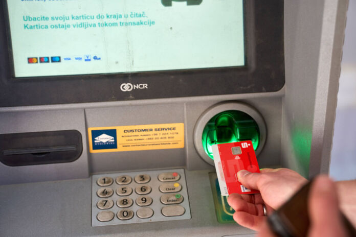 Eine rote Bankkarte wird in den Schlitz beim Automaten gesteckt. Der Bildschirm gibt Anweisungen an den Kunden, wie er den Automaten nutzen kann.