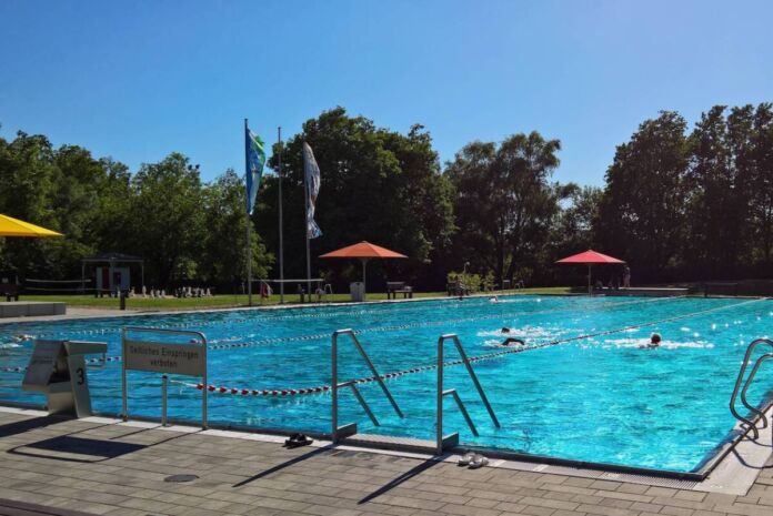 Ein großes Sportbecken mit vier Bahnen in einem Freibad. Einige Besucher schwimmen. Im Hintergrund ist eine grüne saftige Wiese, ein großes Schachbrett, Sonnenschirme und hohe Bäume. Es ist ein Sommertag, der Himmel ist blau. Ein Freibad bei Karlsruhe muss im Juni schließen.