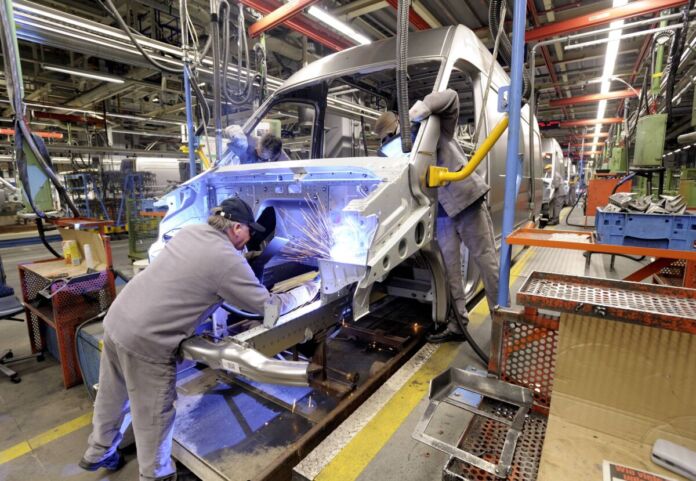 Zwei Männer schweißen an einem großen, silbernen Transporter in einer Fabrik. Die Mitarbeiter tragen graue Arbeitskleidung. Hinter dem Fahrzeug stehen Dutzende weitere.
