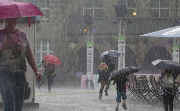 Unwetter mit einer Regenflut in einer Stadt. Über einen Platz laufen viele Menschen mit Regenschirmen. Es regnet sehr stark und der Boden ist bereits mit Wasser bedeckt. Auch die Menschen selbst sind unter ihren Regenschirmen nass und es scheint zudem zu stürmen.  