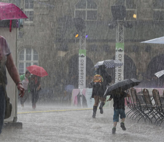 Unwetter mit einer Regenflut in einer Stadt. Über einen Platz laufen viele Menschen mit Regenschirmen. Es regnet sehr stark und der Boden ist bereits mit Wasser bedeckt. Auch die Menschen selbst sind unter ihren Regenschirmen nass und es scheint zudem zu stürmen.  