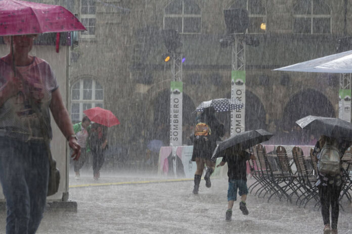 Unwetter mit einer Regenflut in einer Stadt. Über einen Platz laufen viele Menschen mit Regenschirmen. Es regnet sehr stark und der Boden ist bereits mit Wasser bedeckt. Auch die Menschen selbst sind unter ihren Regenschirmen nass und es scheint zudem zu stürmen.  