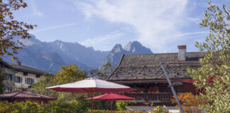 Vor einer wunderschönen Bergkulisse steht eine Wirtschaft, die einen Biergarten mit vielen Draußenplätzen versorgt. Sonnenschirme sind aufgespannt, die Tische noch leer und das Ambiente ist ruhig und das Grundstück mit Pflanzen und Blumen gesäumt.