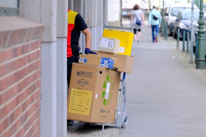 Ein DHL-Bote in gelb-roter Uniform an einer Straße steht mit einem Schubkarren voller großer und mittelgroßer Päckchen und Pakete vor einem Hauseingang und betätigt die Klingel oder will ins Haus eintreten.