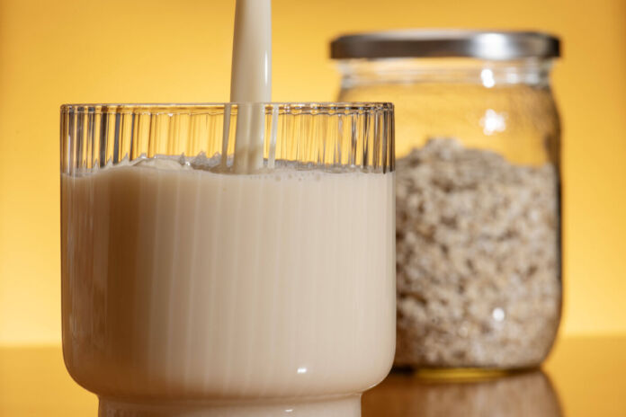 Neben einem Glas voller Hafer vor gelben Hintergrund wird in ein Glas von oben eine Flüssigkeit, wohl Hafermilch, eingegossen. Das Glas ist fast voll und die Milch wirkt dickflüssig und leicht bräunlich, aber noch frisch.