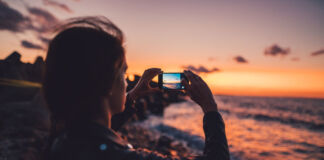 Eine Frau macht am Strand mit der Kamers Ihres Smartphones ein Foto eines wunderschönen Sonnenuntergangs am Strand. Die Wellen fließen über den Sand, während der Himmel rot-orange strahlend leuchtet.