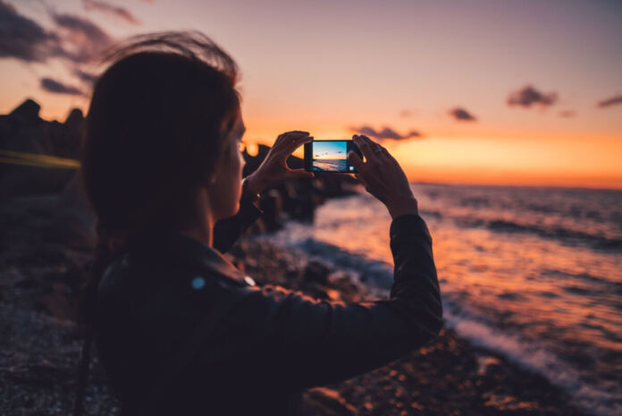 Eine Frau macht am Strand mit der Kamers Ihres Smartphones ein Foto eines wunderschönen Sonnenuntergangs am Strand. Die Wellen fließen über den Sand, während der Himmel rot-orange strahlend leuchtet.