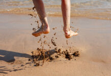 Eine Person hüpft im Sand an einem schönen Strand am Meer. Im Moment der Fotoaufnahme sind ihre Füße in der Luft und es sind Schlammspritzer unter ihr zu sehen.