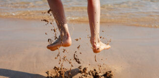 Eine Person hüpft im Sand an einem schönen Strand am Meer. Im Moment der Fotoaufnahme sind ihre Füße in der Luft und es sind Schlammspritzer unter ihr zu sehen.