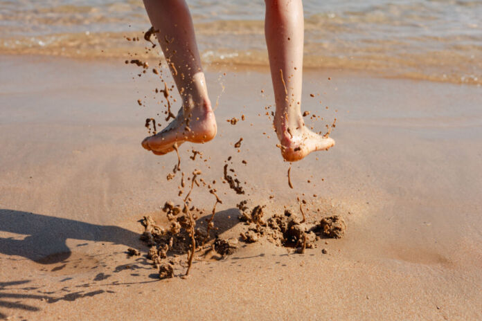 Eine Person hüpft im Sand an einem schönen Strand am Meer. Im Moment der Fotoaufnahme sind ihre Füße in der Luft und es sind Schlammspritzer unter ihr zu sehen.