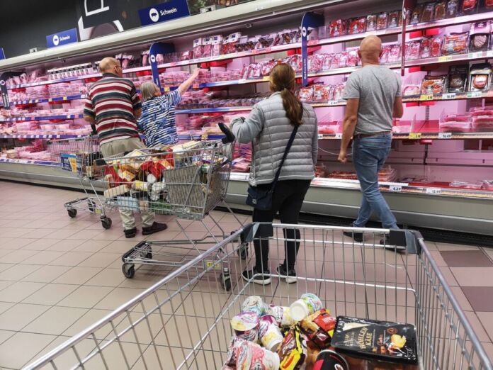 Mehrere Menschen mit Einkaufswagen stehen vor einem Tiefkühlregal mit Fleisch, Wurst und Geflügel in einem Supermarkt oder Discounter und treffen ihre Einkaufwahl.