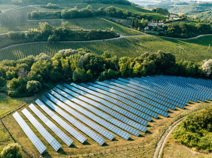 Auf einer Luftaufnahme eines Solarparks auf dem Land sind zahlreiche Solarpanels zu sehen, die umringt von Bäumen mitten in der Landschaft auf einer grünen Wiese in der Nähe eines Dorfes stehen und Strom und Sonnenenergie liefern. Eine Solar-Firma schlittert nun in die Insolvenz.