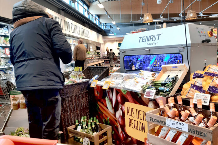 Ein Mann steht in einer dicken Winterjacke in einem Supermarkt oder Discounter direkt vor dem Gemüsestand. Hier kann man erkennen, dass gerade Spargelzeit ist. Verschiedene Sorten von weißem Spargel und auch grünem Spargel sind ausgelegt.