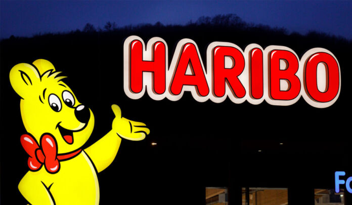Der berühmte Goldbär von Haribo als Leuchtreklame vor einer Fabrik der Firma Haribo. Es ist noch dunkel und es findet offenbar ein Fabrikverkauf für die Süßigkeiten und das Fruchtgummi des Herstellers statt.