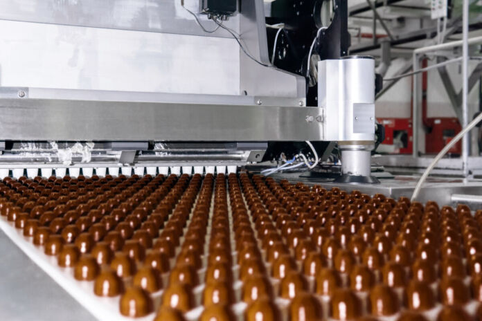 Eine Nahaufnahme eines Förderbandes in einer Süßigkeitenfabrik voll mit Schokopralinen, die beim Durchfahren von einer automatisierten Maschine mit Vollmilchschokolade überzogen werden.