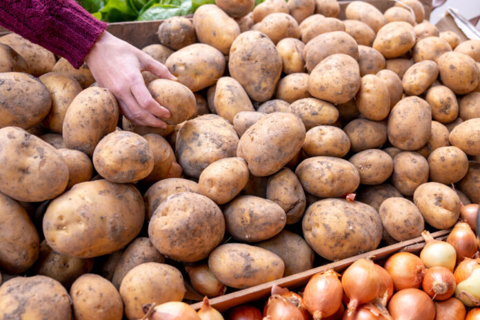 Die Hand einer Frau greift in eine große Holzbox, in der viele Kartoffeln auf einem riesigen Stapel liegen, und nimmt sich eine Kartoffel heraus. Daneben befindet sich eine Box mit Zwiebeln.