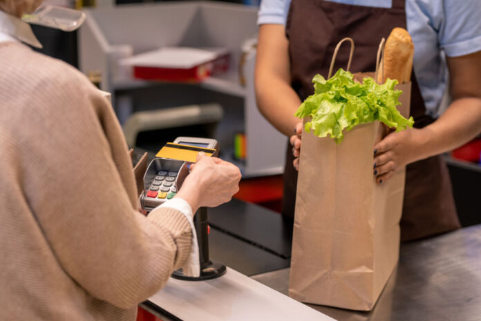 Eine Kundin steht an der Supermarktkasse bei einer Kassiererin. Sie bezahlt, indem sie ihre EC-Karte vor das Lesegerät hält. Die Mitarbeiterin hält die Tüte mit den Einkäufen. Hier gucken Salat und ein Baguette heraus.