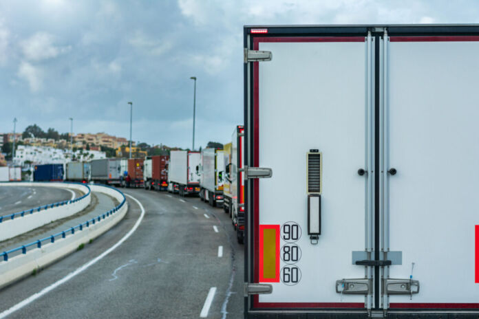 Eine Reihe Lastwagen steht hintereinander auf der Autobahn. Der Stau der LKW könnte von Grenzkontrollen oder dem Zoll herrühren. Im Hintergrund ist eine südlich aussehende Stadt zu erkennen.