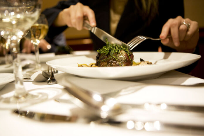Eine Person sitzt an einem Tisch in einem guten Restaurant oder Café. Er oder sie isst mit Messer und Gabel eine Delikatesse, vielleicht Fleisch, Kartoffeln und frisches Gemüse.