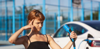 Ein schönes junges Mädchen hält ein Ladekabel vor einem weißen E-Auto. Sie verzieht das Gesicht und hält einen Finger an die Stirn, um Zweifel oder Unwissenheit auszudrücken. Anscheinend gibt es Probleme beim Aufladen des Stromers.