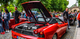 Ein roter Ferrari von hinten. Der Sportwagen steht am Straßenrand. Sein Heck ist geöffnet und offenbart den riesigen V12-Motor. Der Rennwagen ist ein Cabrio.