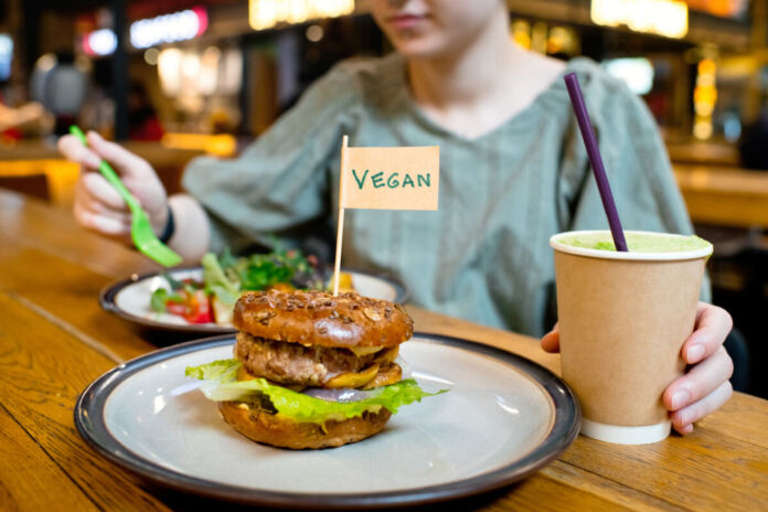 Eine Frau hat in einem veganen Restaurant an einen Burger bestellt. Dazu hat sie ein Getränk und einen kleinen Salat, den sie gerade isst.