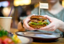 Eine Person oder ein Gast hält einen veganen Burger mit beiden Händen. Oben auf dem Burger-Patty ist ein kleines Fähnchen eingesteckt mit der Aufschrift "vegan".