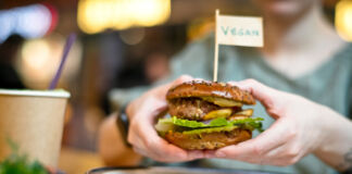 Eine Person oder ein Gast hält einen veganen Burger mit beiden Händen. Oben auf dem ausgiebig belegten Burger samt Burger-Patty ist ein kleines Fähnchen eingesteckt mit der Aufschrift "vegan".