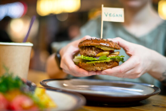 Eine Person oder ein Gast hält einen veganen Burger mit beiden Händen. Oben auf dem ausgiebig belegten Burger samt Burger-Patty ist ein kleines Fähnchen eingesteckt mit der Aufschrift "vegan".